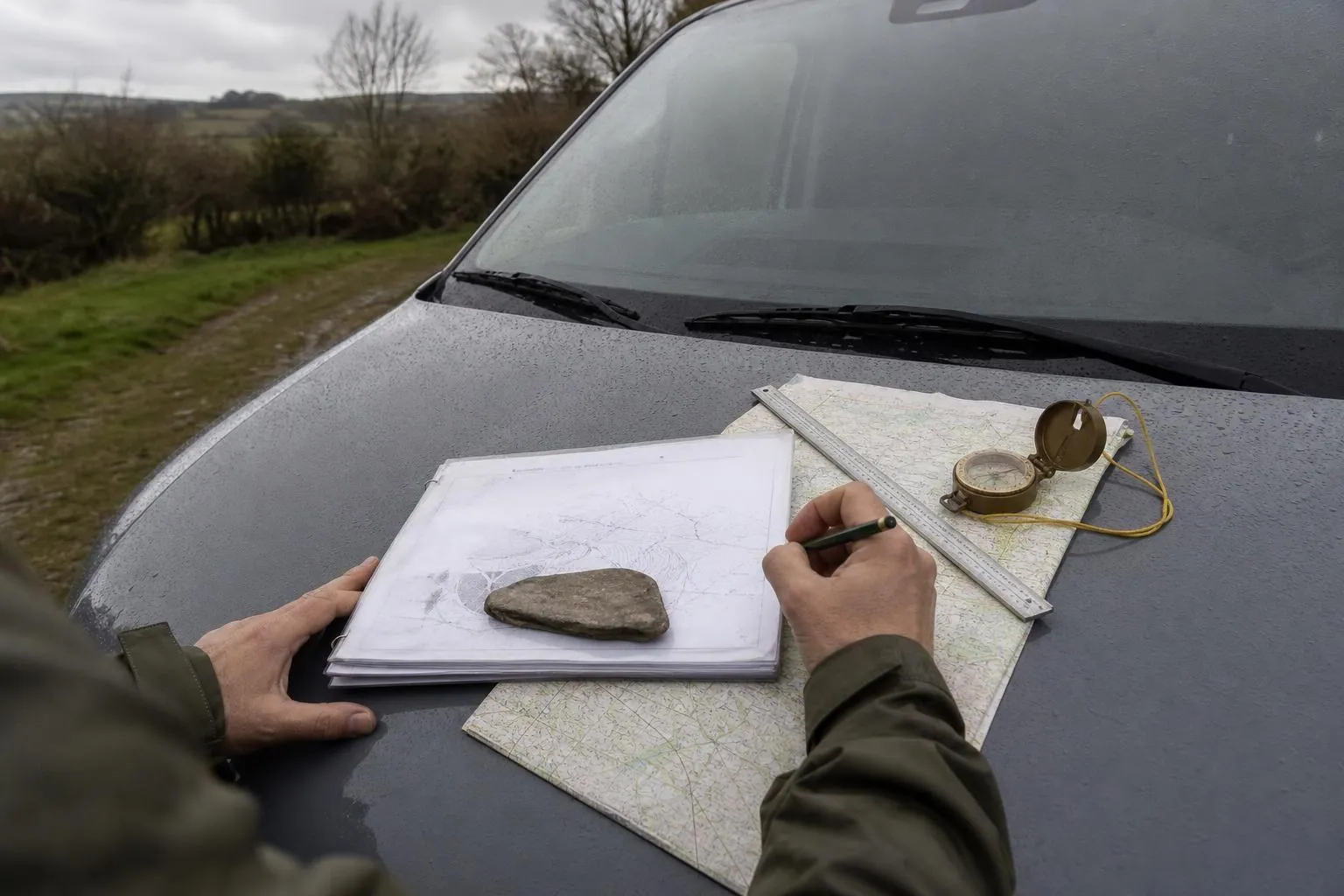 Liasse de fiches de terrain agrafees, carte topographique pliee et boussole posees sur le capot mouille d un fourgon en bord de chemin agricole.