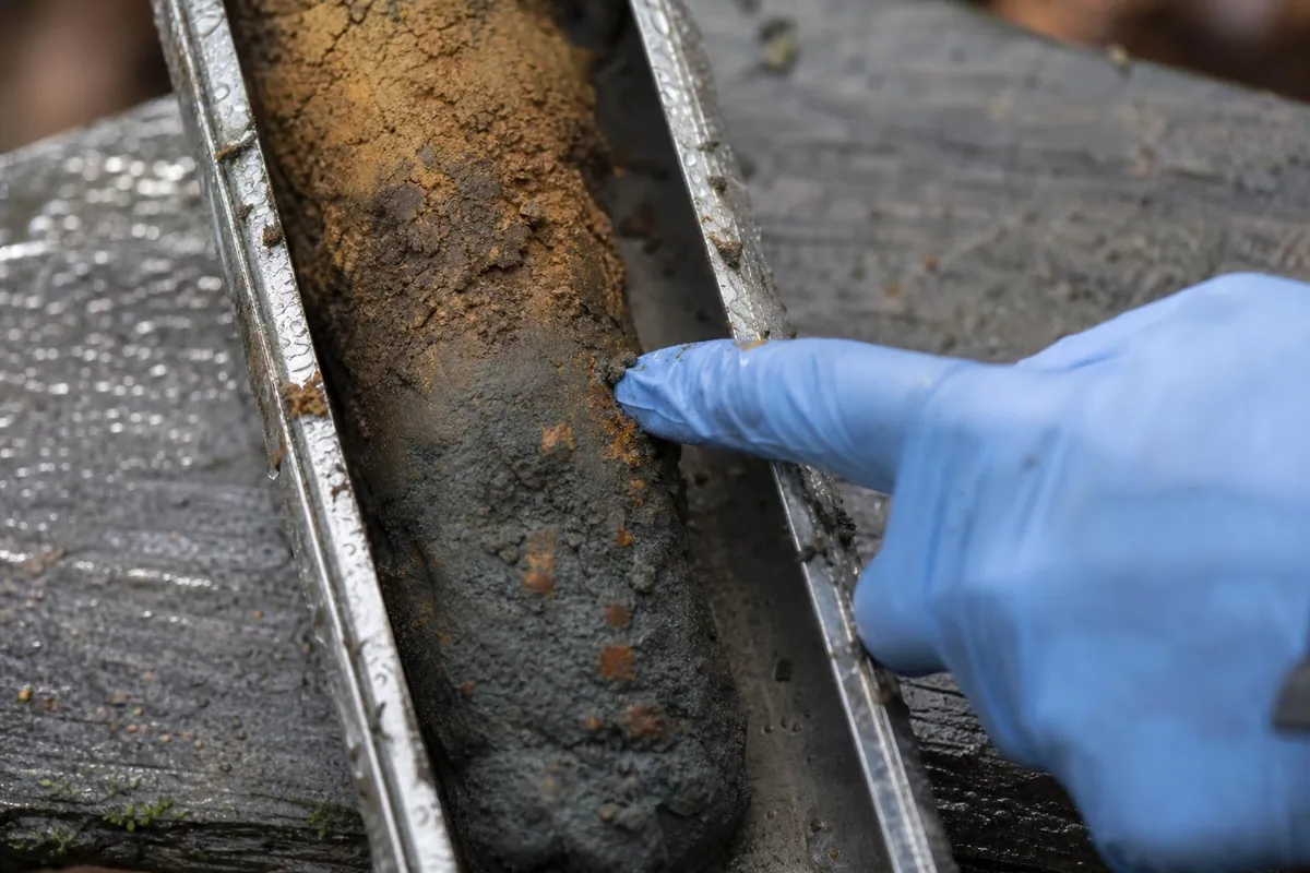 Gros plan d une carotte de sol dans la goulotte metallique d une tariere, avec des taches rouille sur un fond gris bleute et des mains gantees en arriere plan